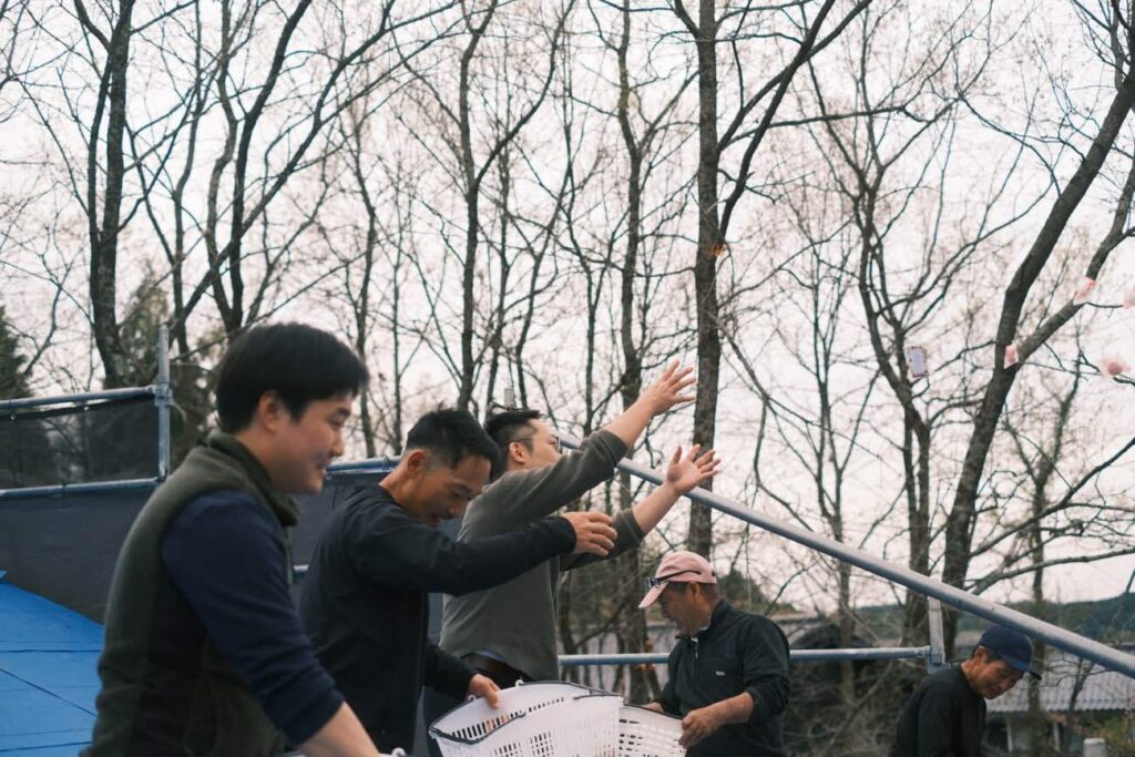 A group of five men outdoors among bare trees, some with arms raised, one holding a white basket, as if celebrating or unloading equipment on a cloudy day.