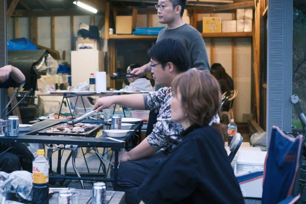 People gather around a grill in a backyard cooking meat and chatting with drinks on the table nearby