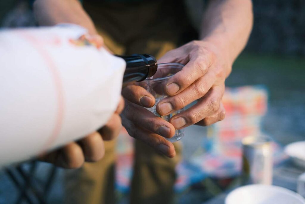 Person pours a drink from a dark bottle into a clear plastic cup at an outdoor picnic with hands steady over a table covered in colorful items
